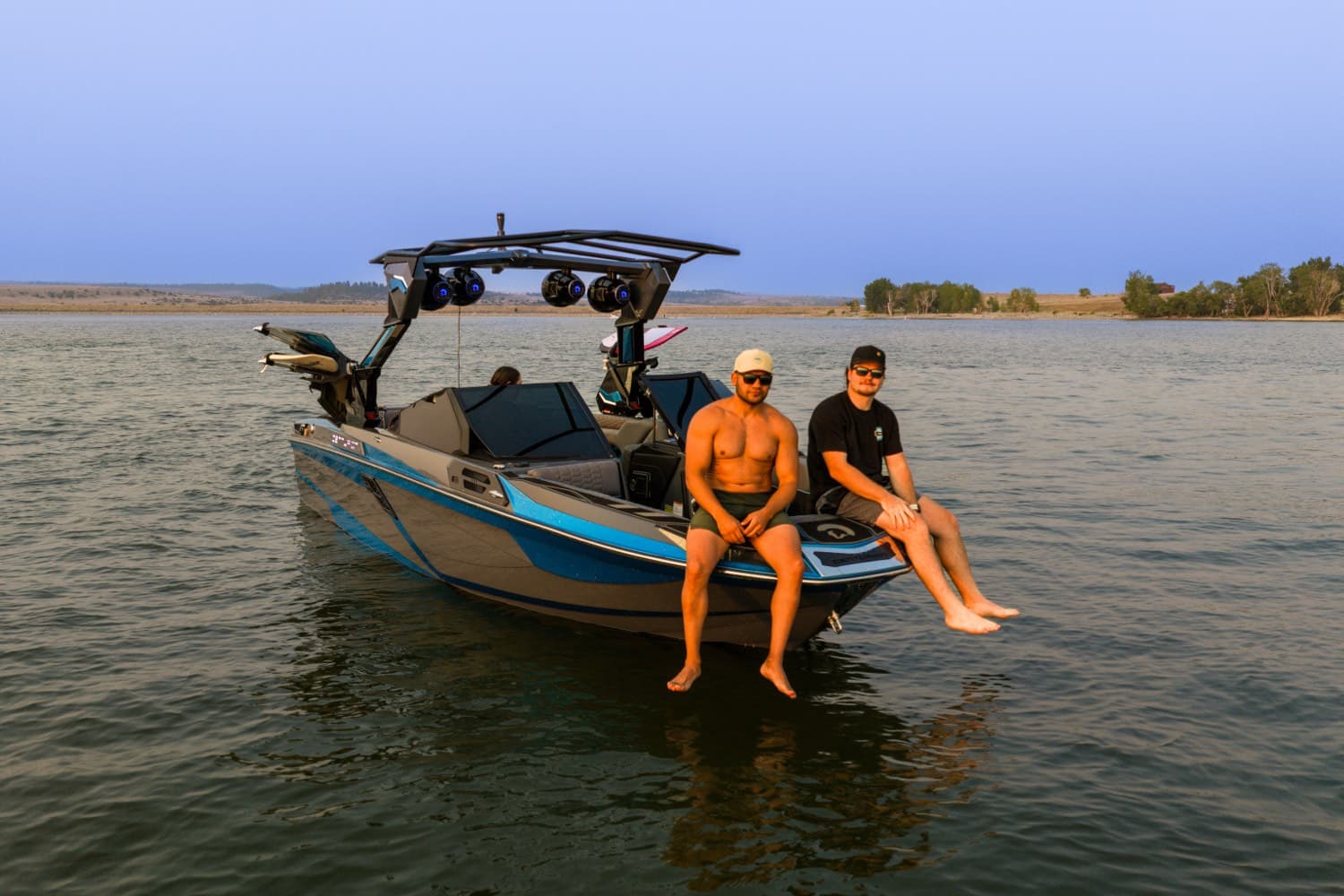 Two people sitting on the transom of a wake boat at dusk, feet in the water.